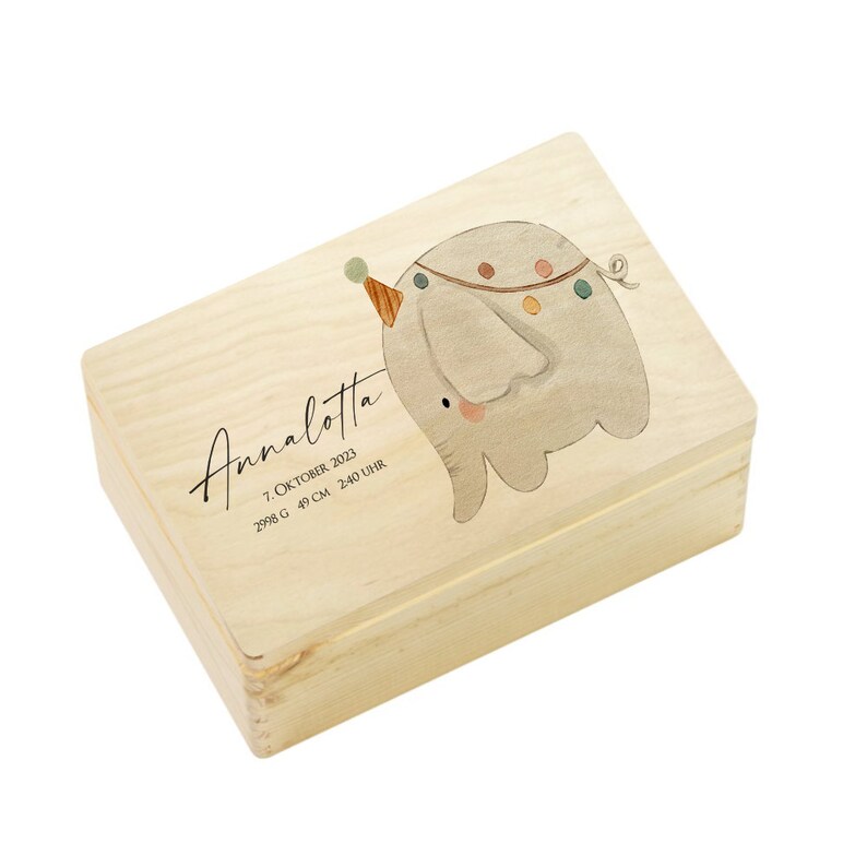 May include: A wooden keepsake box with a light wood finish. The box features a watercolour illustration of a grey elephant wearing a party hat and a string of colourful lights. The text "Annalotta" is written in cursive script, along with the date "7. Oktober 2023", "2998 g", "49 cm", and "2:40 Uhr".