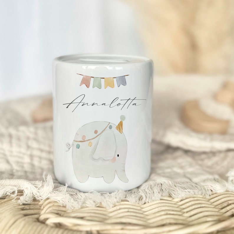 May include: White ceramic piggy bank with a cartoon elephant wearing a party hat and holding colorful flags. The piggy bank is decorated with the name "Annalotta".