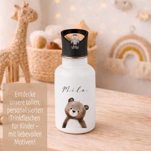 May include: White water bottle with a black lid, featuring a cartoon bear illustration and the name "Milo" in script. The bottle has a flip-top cap and is designed for children. The image also includes text in German.