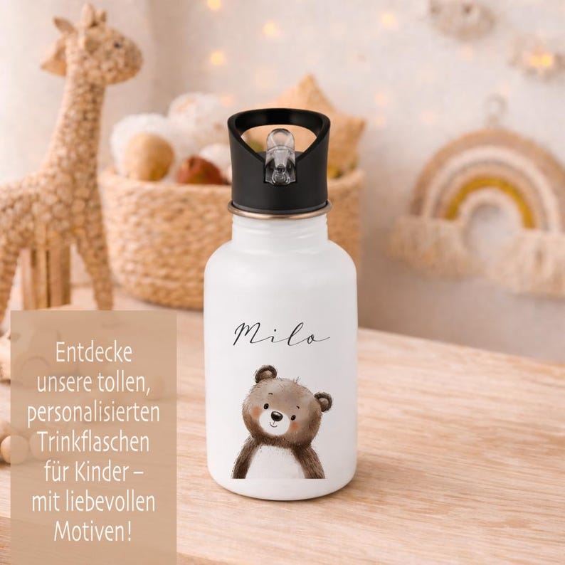 May include: White water bottle with a black lid, featuring a cartoon bear illustration and the name "Milo" in script. The bottle has a flip-top cap and is designed for children. The image also includes text in German.