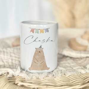 May include: White ceramic piggy bank with a brown bear wearing a party hat and colorful bunting. The name "Chaska" is written on the front.