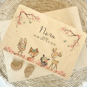 May include: Wooden plaque with a floral design and a name, Nora, with birth details: 25.04.2023, 18:37 Uhr, 4220 g, 56 cm, 38.5 cm. The plaque features a bunny, fox, raccoon, and deer.