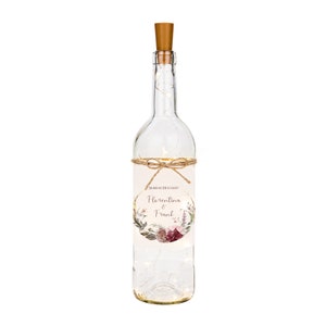 Könnte beinhalten: Eine klare Glasflasche mit einem Korkverschluss und einer Lichterkette aus weißen Lichtern im Inneren. Die Flasche hat ein Etikett mit einem Blumendesign und dem Text "Florentina & Frank" in einem Kreis.