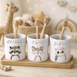 Op de afbeelding: Drie witte keramische tandenborstelhouders met illustraties van een wasbeer, vos en uil. Elke houder heeft een naam: Oliver, Ezra en Sofia. De houders staan op een houten dienblad, elk met een houten tandenborstel.