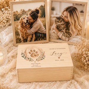 May include: A rectangular light-colored wooden box featuring a laser-engraved design of a paw print surrounded by a wreath. The text reads "GELIEBT & UNVERGESSEN", "Lucy", "2014 - 2026", "MEIN BESTER", "Freund", and "AUF 4 PFOTEN" on the lid.