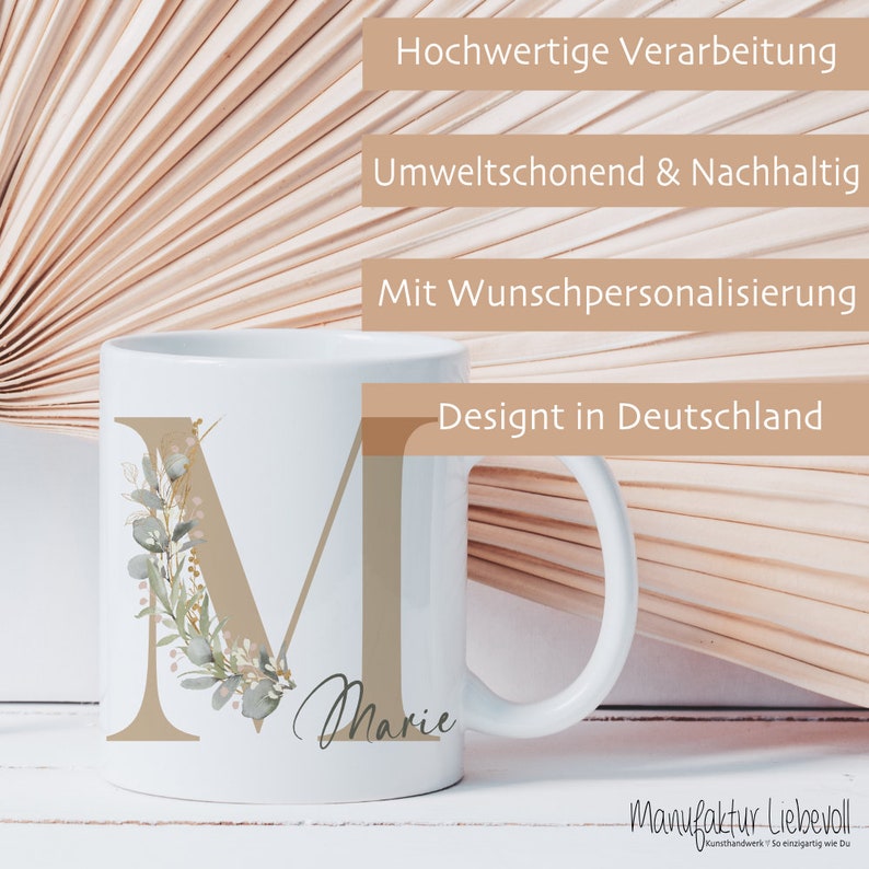 Puede incluir: Taza de cer&aacute;mica blanca con una letra M marr&oacute;n decorada con hojas verdes y flores rosas. El nombre Marie est&aacute; escrito en cursiva debajo de la letra. La taza est&aacute; sobre un fondo de un abanico de madera.