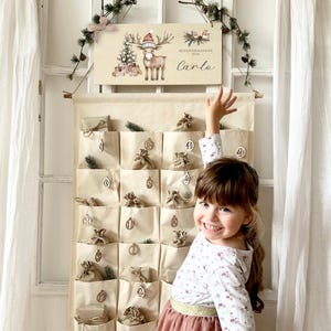 Può includere: Un calendario dell'Avvento in tessuto beige con piccole tasche, ognuna contenente un piccolo regalo. Un cartello di legno sopra raffigura una renna con un cappello da Babbo Natale e il nome "Carlo". Il calendario è decorato con piccoli ramoscelli verdi.
