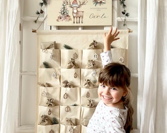 May include: A beige fabric advent calendar with small pockets, each holding a small gift. A wooden sign above features a reindeer wearing a Santa hat and the name "Carlo". The calendar is decorated with small sprigs of greenery.