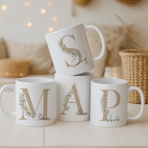 Op de afbeelding: Witte keramische mokken met grote, bruine letters: M, A, P en S. Elke letter is versierd met een bloemenontwerp en een naam. De mok met de letter "S" heeft de naam "Sunny" erop geschreven. De mokken staan op een houten oppervlak.