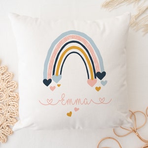 May include: A white pillow with a rainbow design in shades of blue, pink, yellow, and black. The rainbow has hearts hanging from it in pink and blue. The name "Emma" is written in pink cursive with hearts on either side.