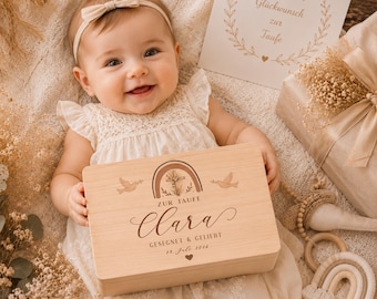 May include: A smiling baby wearing a cream-colored dress and headband holds a wooden box. The box features a rainbow design, doves, and the name "Clara" with text below. A card with text and a gift box are in the background.