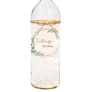 May include: A clear glass bottle with a white label that says "Lieblings MAMA" in a gold geometric frame with green eucalyptus leaves. The bottle is decorated with a string of lights and a brown twine bow.