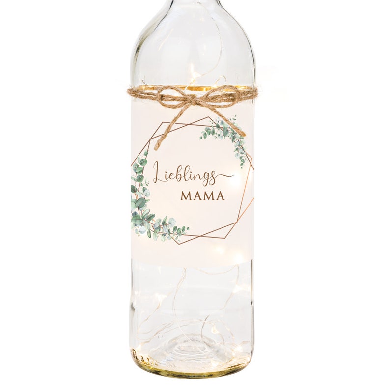 May include: A clear glass bottle with a white label that says "Lieblings MAMA" in a gold geometric frame with green eucalyptus leaves. The bottle is decorated with a string of lights and a brown twine bow.