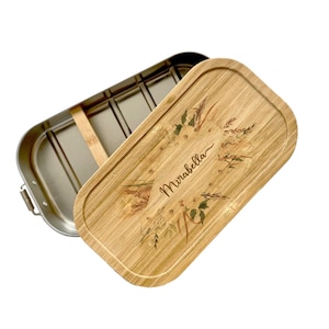 Op de afbeelding: Een lunchbox van roestvrij staal met een bamboe deksel. Het deksel heeft een bloemenmotief en de naam "Mirabella" erop.