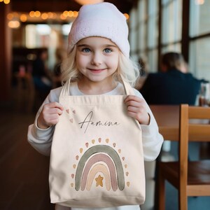 Peut inclure: Un sac fourre-tout en toile blanche avec un motif arc-en-ciel et le nom "Amina" imprim&eacute; dessus. L'arc-en-ciel est dans des tons de rose, orange et vert avec une &eacute;toile dor&eacute;e au centre.