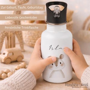 May include: A white water bottle with a black lid, held by a child. The bottle features the name "Isla" in script, a cartoon squirrel, and text in German. The bottle is a personalized gift idea for a baby shower or birthday.
