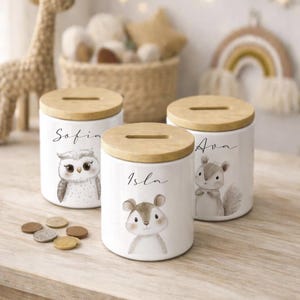 Puede incluir: Tres huchas de cer&aacute;mica blanca con tapas de madera. Cada hucha presenta una ilustraci&oacute;n de un animal del bosque: un b&uacute;ho, un rat&oacute;n y una ardilla. Los nombres "Sofia", "Isla" y "Ava" est&aacute;n escritos en las huchas.