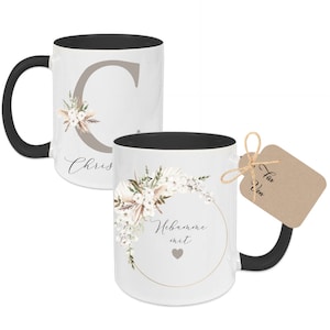 Op de afbeelding: Twee zwart-witte koffiemokken met een bloemenontwerp. Een mok heeft een grote letter C en de tekst "Chris" erop. De andere mok heeft een bloemenkrans met de tekst "Hebamme mit" en een hart. Een bruin cadeau-etiket met de tekst "Für den" is bevestigd aan de tweede mok.