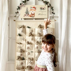Puede incluir: Calendario de Adviento de tela beige con pequeños bolsillos, cada uno con un regalo envuelto. Un letrero de madera en la parte superior presenta un zorro con un gorro de Papá Noel y el texto "Adventskalender von Petra."