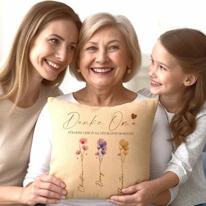 May include: A beige throw pillow with the words "Danke Oma" and "F&Uuml;R DEINE LIEBE IN ALL DEN KLEINEN MOMENTEN" printed in brown script. The pillow features three floral illustrations with names below them. The pillow is held by a woman.