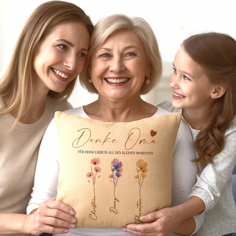 May include: A beige throw pillow with the words "Danke Oma" and "F&Uuml;R DEINE LIEBE IN ALL DEN KLEINEN MOMENTEN" printed in brown script. The pillow features three floral illustrations with names below them. The pillow is held by a woman.