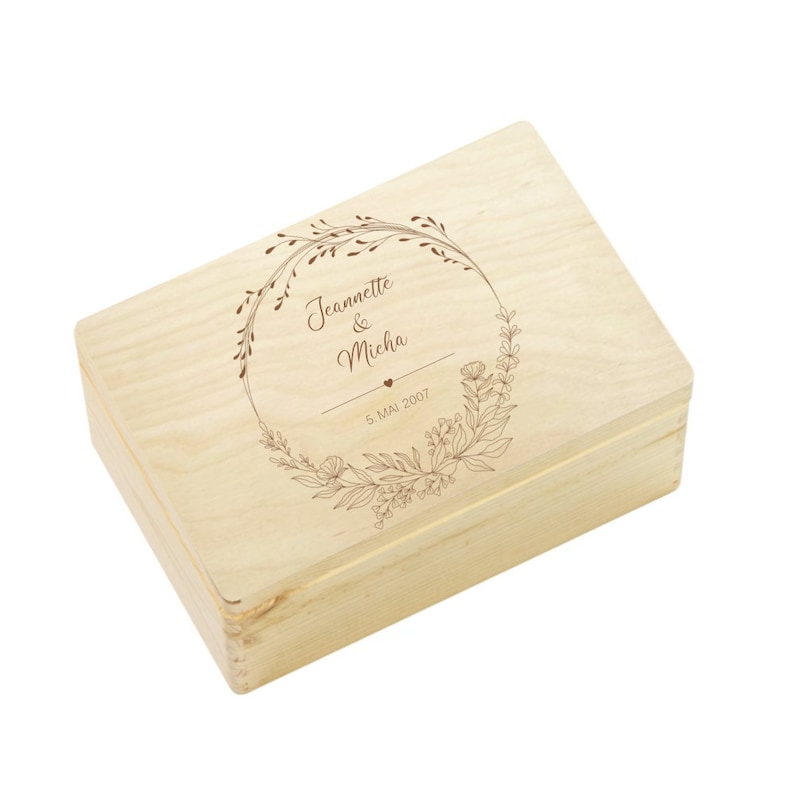 May include: A light wood box with a floral wreath design engraved on the lid. The wreath includes the names "Jeannette & Micha" and the date "5. MAI 2007".