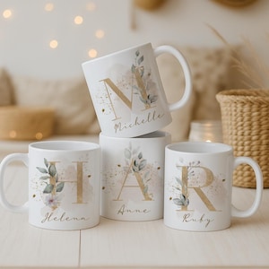 Può includere: Quattro tazze in ceramica bianca con scritte dorate e dettagli floreali. Ogni tazza presenta un'iniziale e un nome diversi: "N Michelle", "H Helena", "A Anne" e "R Ruby". Le tazze hanno un design classico ed elegante.