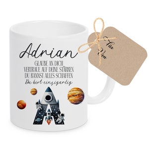 Könnte beinhalten: Weiße Keramiktasse mit einem Raketen- und Planetenmotiv. Die Tasse trägt den Namen "Adrian" und den Text "GLAUBE AN DICH, VERTRAUE AUF DEINE STÄRKEN, DU KANNST ALLES SCHAFFEN, Du bist einzigartig". Ein braunes Geschenkanhänger mit dem Text "Für Von" ist an der Tasse befestigt.
