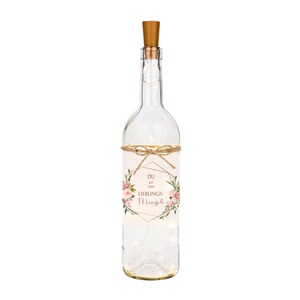 May include: A clear glass bottle with a cork top and a label that reads "Du bist mein Lieblings-Mensch". The bottle is decorated with pink flowers and a gold geometric frame. The bottle is filled with white fairy lights.