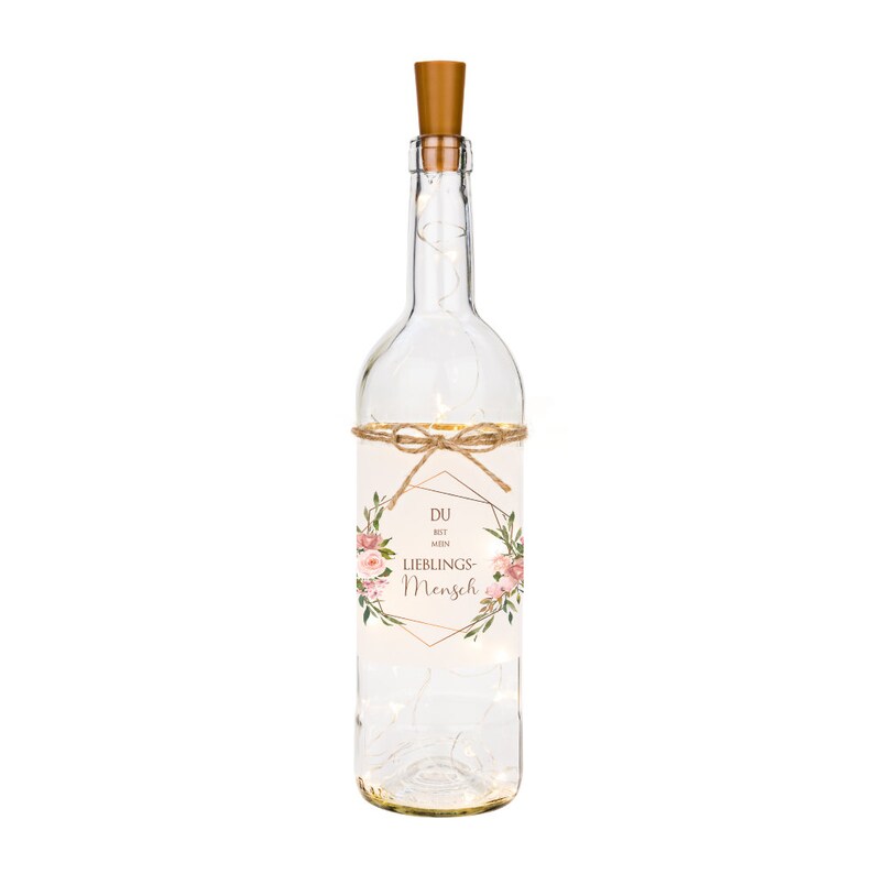 May include: A clear glass bottle with a cork top and a label that reads "Du bist mein Lieblings-Mensch". The bottle is decorated with pink flowers and a gold geometric frame. The bottle is filled with white fairy lights.