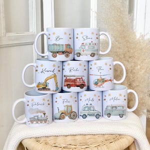 Puede incluir: Un juego de diez tazas de cerámica blanca con diferentes diseños de vehículos y nombres impresos en ellas. Las tazas presentan un camión, una ambulancia, una excavadora, un camión de bomberos, un helicóptero, un tren, un tractor, un coche de policía y una furgoneta.