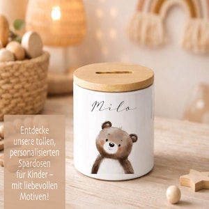Puede incluir: Hucha de cer&aacute;mica blanca con tapa de madera. El banco presenta una ilustraci&oacute;n de oso de dibujos animados y el nombre "Milo" en escritura cursiva. El texto "Entdecke unsere tollen, personalisierten Spardosen f&uuml;r Kinder - mit liebevollen Motiven!" tambi&eacute;n es visible.