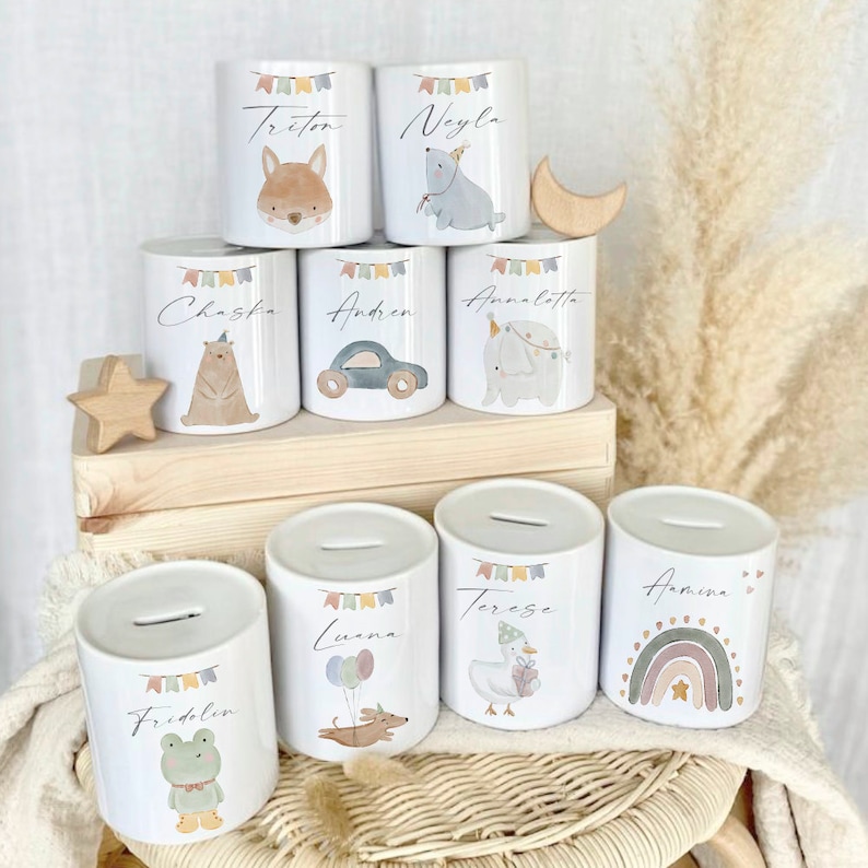 May include: Set of ten white ceramic piggy banks with colorful designs and personalized names. Each bank features a different animal or object, including a fox, a bear, a car, an elephant, a dog, a goose, a frog, and a rainbow.
