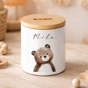 Puede incluir: Hucha de cer&aacute;mica blanca con tapa de madera. El banco presenta una ilustraci&oacute;n de oso de dibujos animados y el nombre "Milo" en escritura cursiva. El oso es marr&oacute;n con una cara marr&oacute;n claro y una peque&ntilde;a nariz negra. La tapa tiene una ranura para insertar monedas.