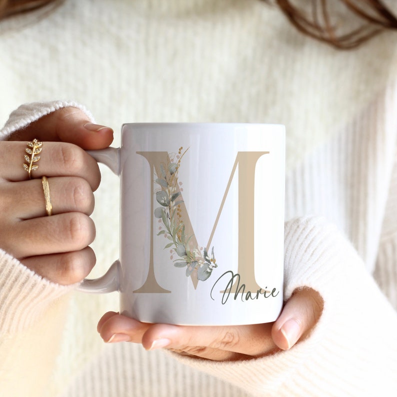 Puede incluir: Taza de cer&aacute;mica blanca con una letra M beige decorada con un dise&ntilde;o floral en tonos verdes y marrones. El nombre "Marie" est&aacute; escrito debajo de la letra.