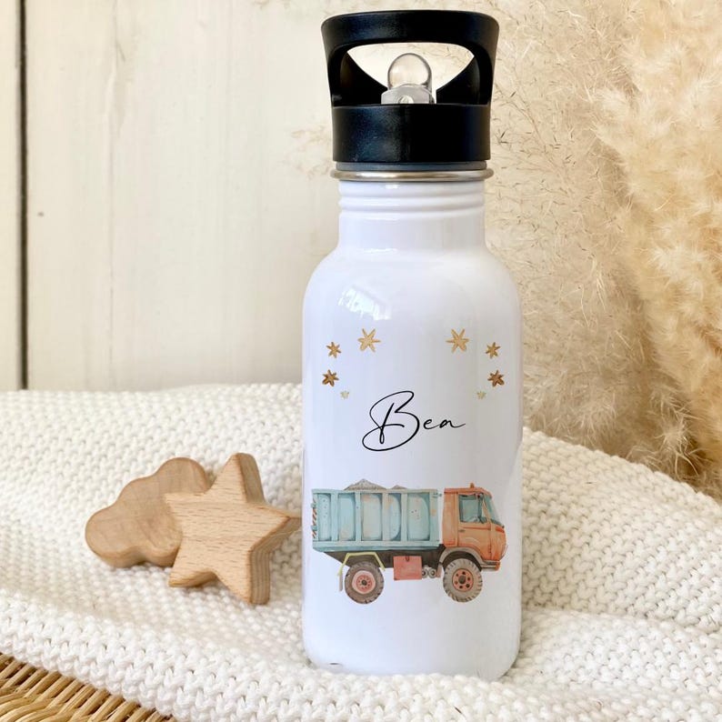 May include: White stainless steel water bottle with a black lid and a handle. The bottle has a colorful illustration of a dump truck and the name "Bea" written in cursive. The bottle is decorated with gold stars.
