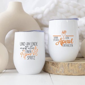 May include: Two white stemless wine glasses with lids. One glass has the text "UND AM ENDE ergibt alles EINEN Aperol SPRITZ" in black and orange. The other glass has the text "KANN SPUREN VON Aperol ENTHALTEN" in black and orange.