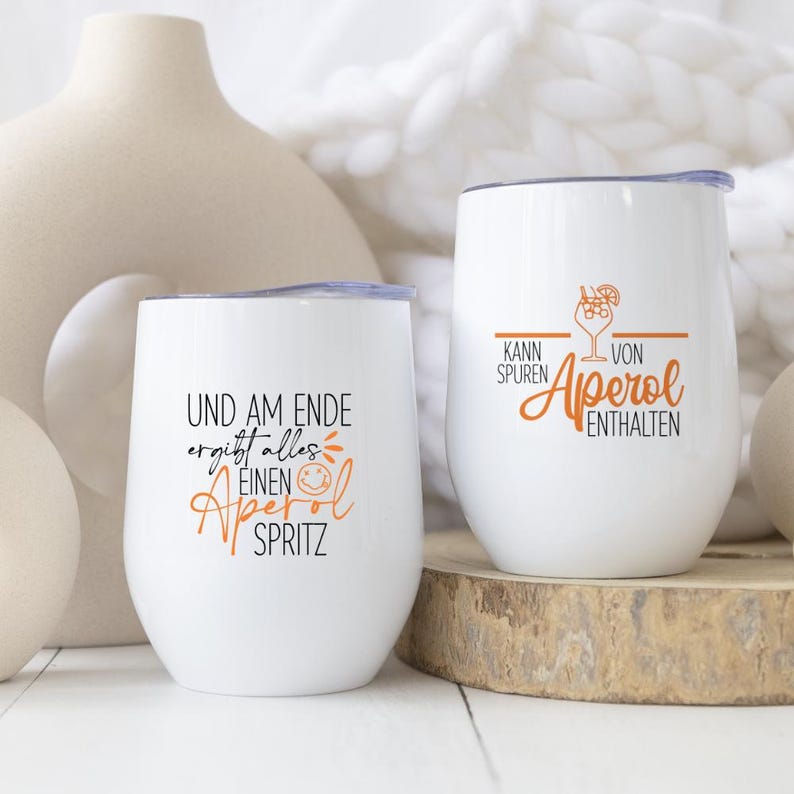 May include: Two white stemless wine glasses with lids. One glass has the text "UND AM ENDE ergibt alles EINEN Aperol SPRITZ" in black and orange. The other glass has the text "KANN SPUREN VON Aperol ENTHALTEN" in black and orange.