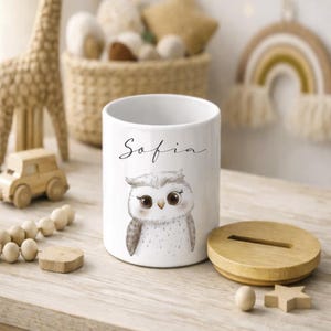 Puede incluir: Hucha de cer&aacute;mica blanca con tapa de madera, con una ilustraci&oacute;n de un b&uacute;ho y el nombre "Sofia". La hucha cil&iacute;ndrica est&aacute; sobre una superficie de madera clara, con otros juguetes de madera en el fondo.