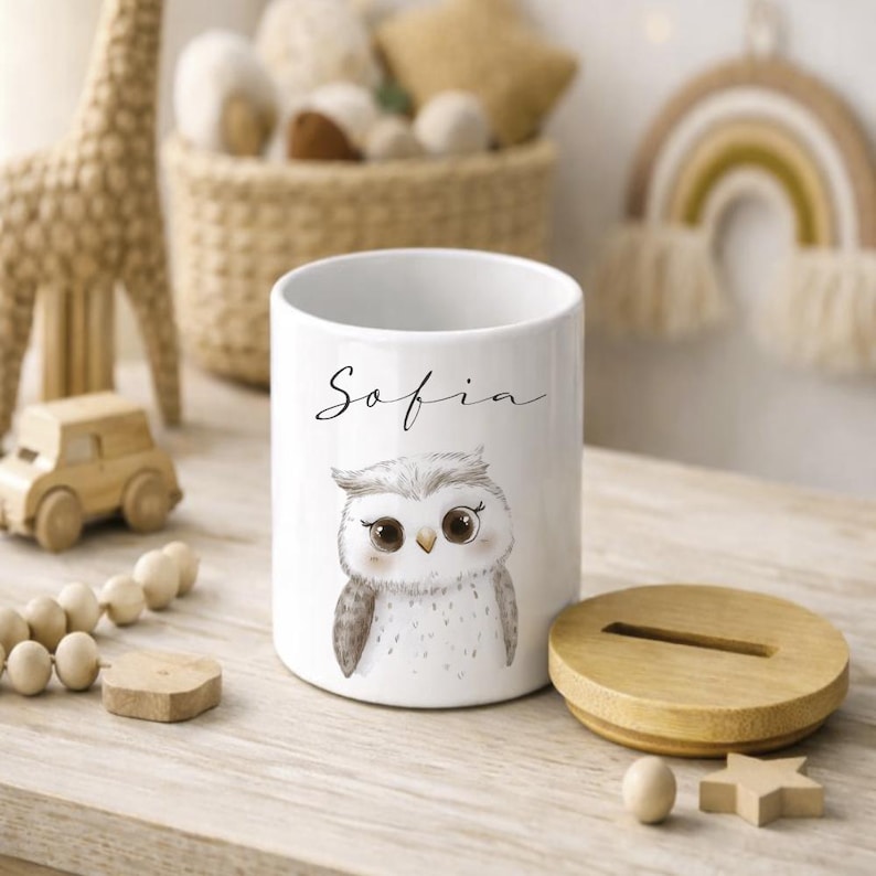 Puede incluir: Hucha de cer&aacute;mica blanca con tapa de madera, con una ilustraci&oacute;n de un b&uacute;ho y el nombre "Sofia". La hucha cil&iacute;ndrica est&aacute; sobre una superficie de madera clara, con otros juguetes de madera en el fondo.