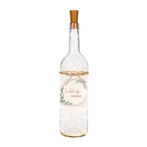 May include: A clear glass bottle with a cork top and a label that says "Lieblings MAMA" in a gold geometric frame with green leaves. The bottle is decorated with a string of lights.