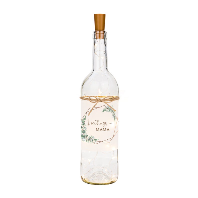 May include: A clear glass bottle with a cork top and a label that says "Lieblings MAMA" in a gold geometric frame with green leaves. The bottle is decorated with a string of lights.
