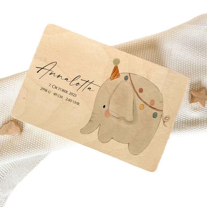 May include: A wooden birth announcement plaque with a hand-painted illustration of a grey elephant wearing a party hat. The plaque reads "Annalotta 7. Oktober 2023 2998 G 49 CM 2:40 UHR".