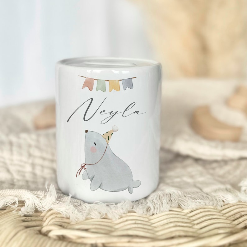 May include: White ceramic piggy bank with a cartoon seal wearing a party hat and a red ribbon. The piggy bank has a colorful banner with the name "Neyla" written on it.