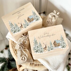 Op de afbeelding: Twee houten adventskalenderdozen met winterse taferelen van besneeuwde bomen, een hut en een hert. De dozen hebben tekst. Kleine jute zakken met houten ornamenten zijn ook aanwezig.