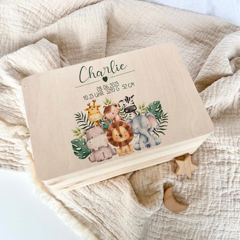 Op de afbeelding: Een houten bewaardoos met een lichte houtkleur. De doos is versierd met een jungledieren thema en de naam "Charlie" met de datum "08.06.2023" en de tijd "10.23 Uhr" samen met het gewicht "3210 g" en de lengte "52 cm".