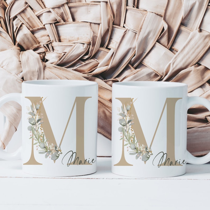 Puede incluir: Dos tazas de cer&aacute;mica blanca con una letra "M" dorada y un dise&ntilde;o floral. Las tazas tienen el nombre "Marie" escrito debajo de la letra.