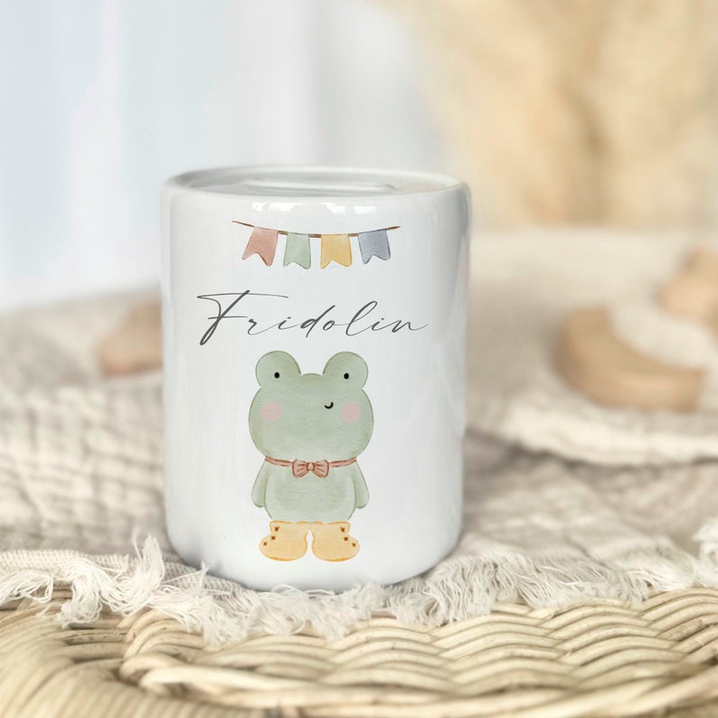May include: White ceramic piggy bank with a green frog wearing a bow tie and yellow boots. The piggy bank has a string of colorful flags and the name "Fridolin" written on it.