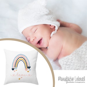May include: A white pillow with a rainbow design and the name "Emma" printed in pink. The rainbow is made of blue, pink, and yellow stripes with small pink hearts falling from it. The pillow is on a white surface.