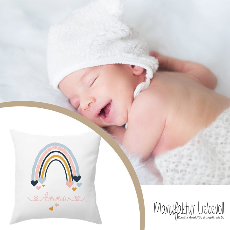May include: A white pillow with a rainbow design and the name "Emma" printed in pink. The rainbow is made of blue, pink, and yellow stripes with small pink hearts falling from it. The pillow is on a white surface.